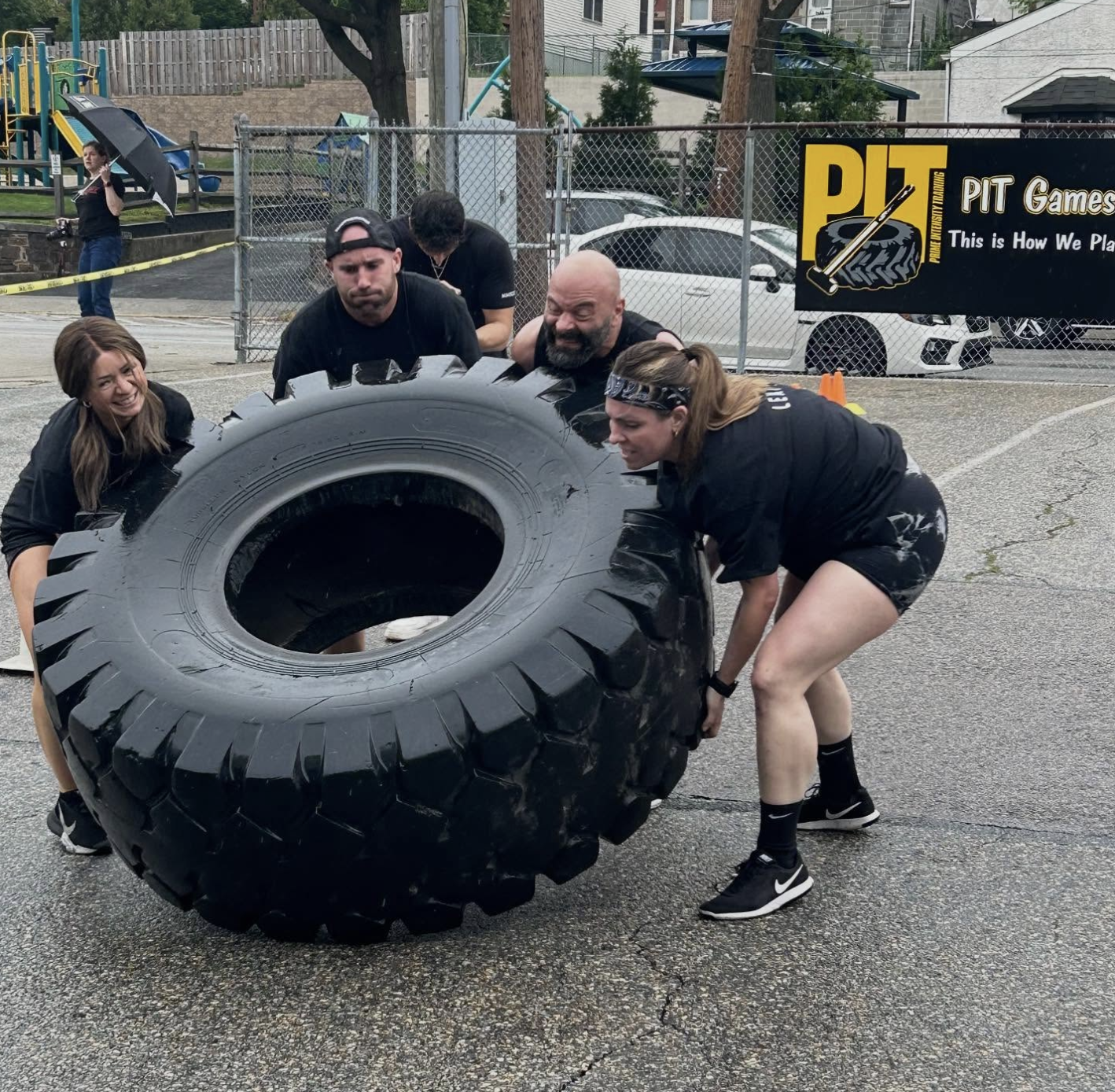 Four people working together to lift and flip a large tractor tire during an outdoor fitness challenge at PIT Gym, with determination on their faces and spectators watching in the background.