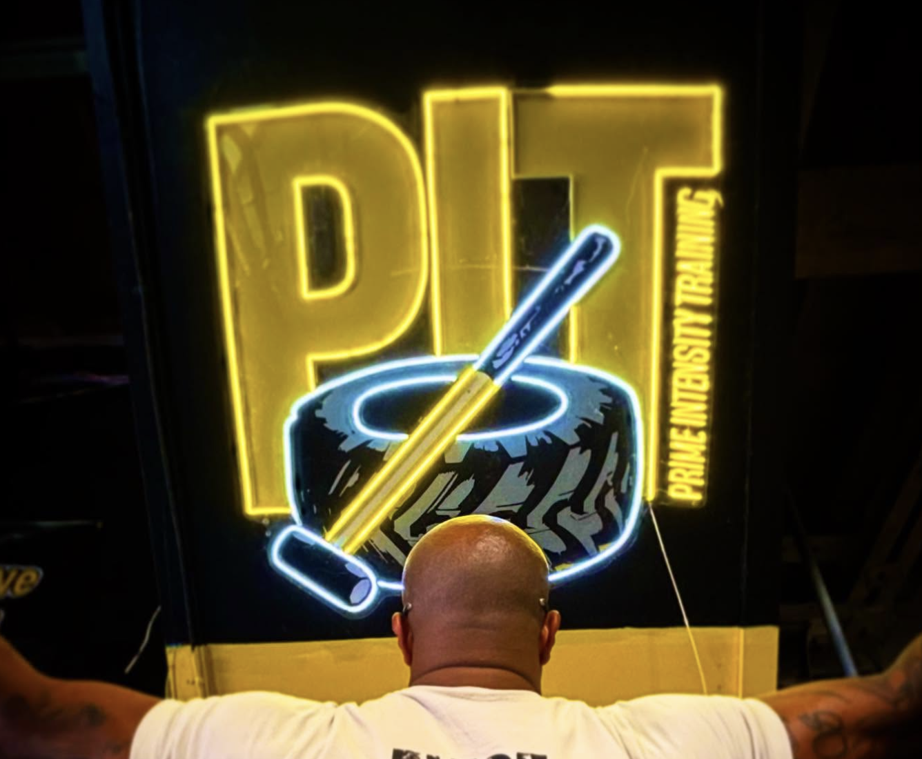 Man facing a bright neon “PIT – Prime Intensity Training” sign inside a gym, wearing a white shirt that reads “EXIST ABOVE ALL LIMITS