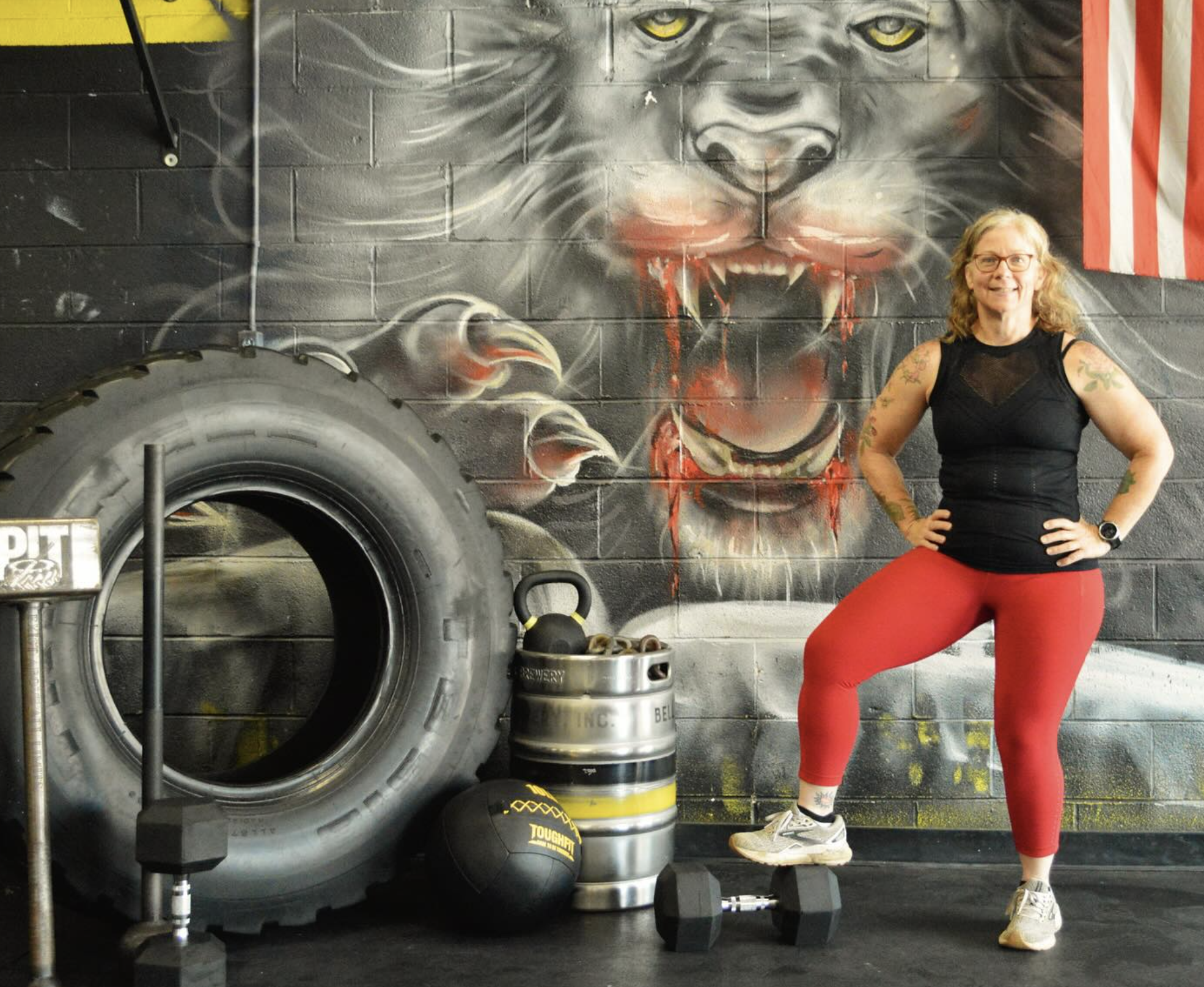 Woman posing confidently in front of a lion mural and American flag at Prime Intensity Training gym, standing beside large tires, weights, and fitness equipment